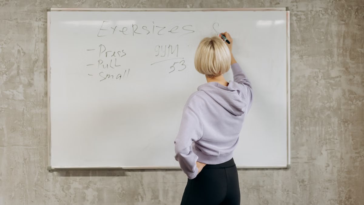 Female writing workout schedule on whiteboard in gym setting.
